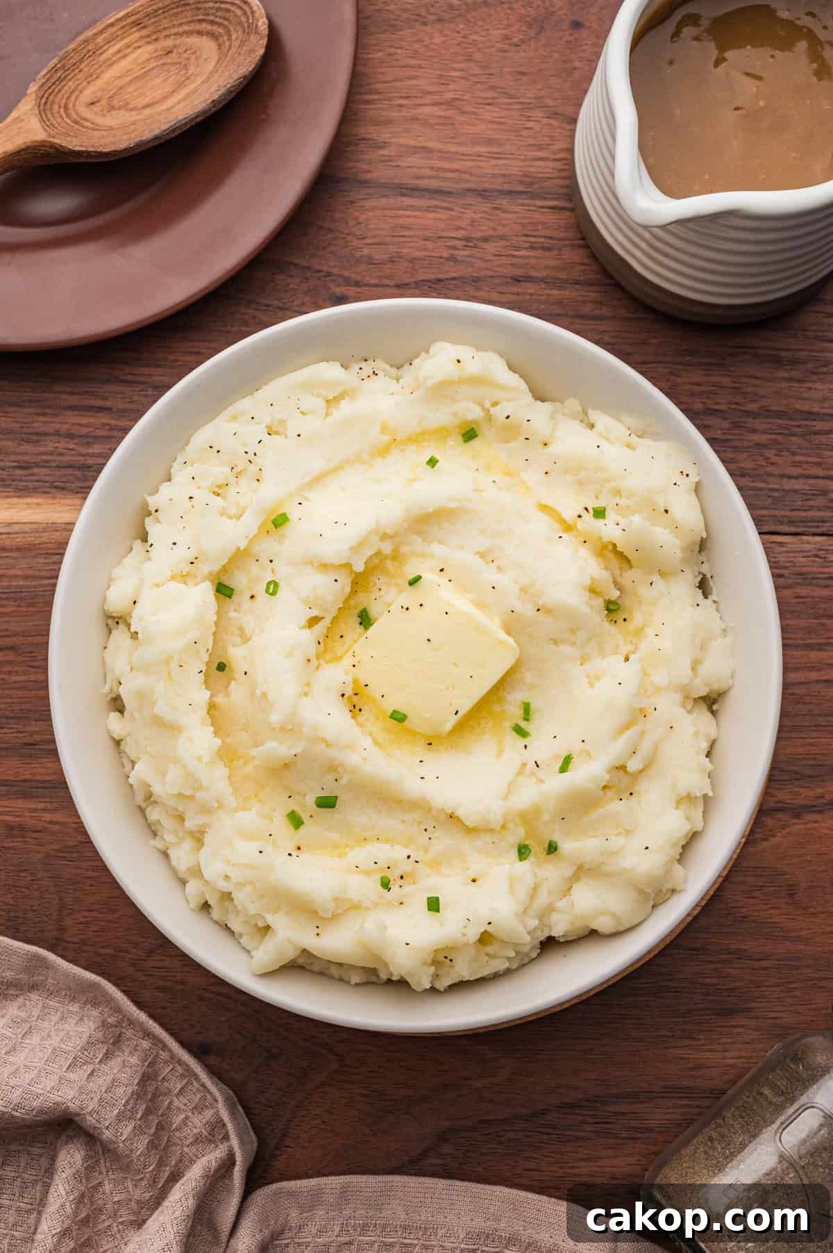 Classic Old-Fashioned Mashed Potatoes Recipe for Creamy Comfort 7 Overhead shot of a bowl of old fashioned mashed potatoes topped with butter, pepper, and chives. The bowl is on a wooden table surrounded by a gravy boat, pepper shaker, wooden spoon, and beige napkins.
