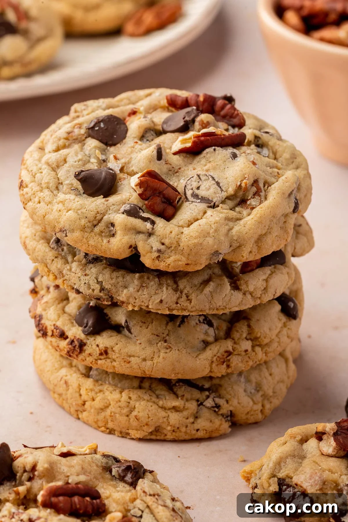 A stack of four gluten free pecan chocolate chip cookies on a pink table with additional cookies and toasted pecans in the background.
