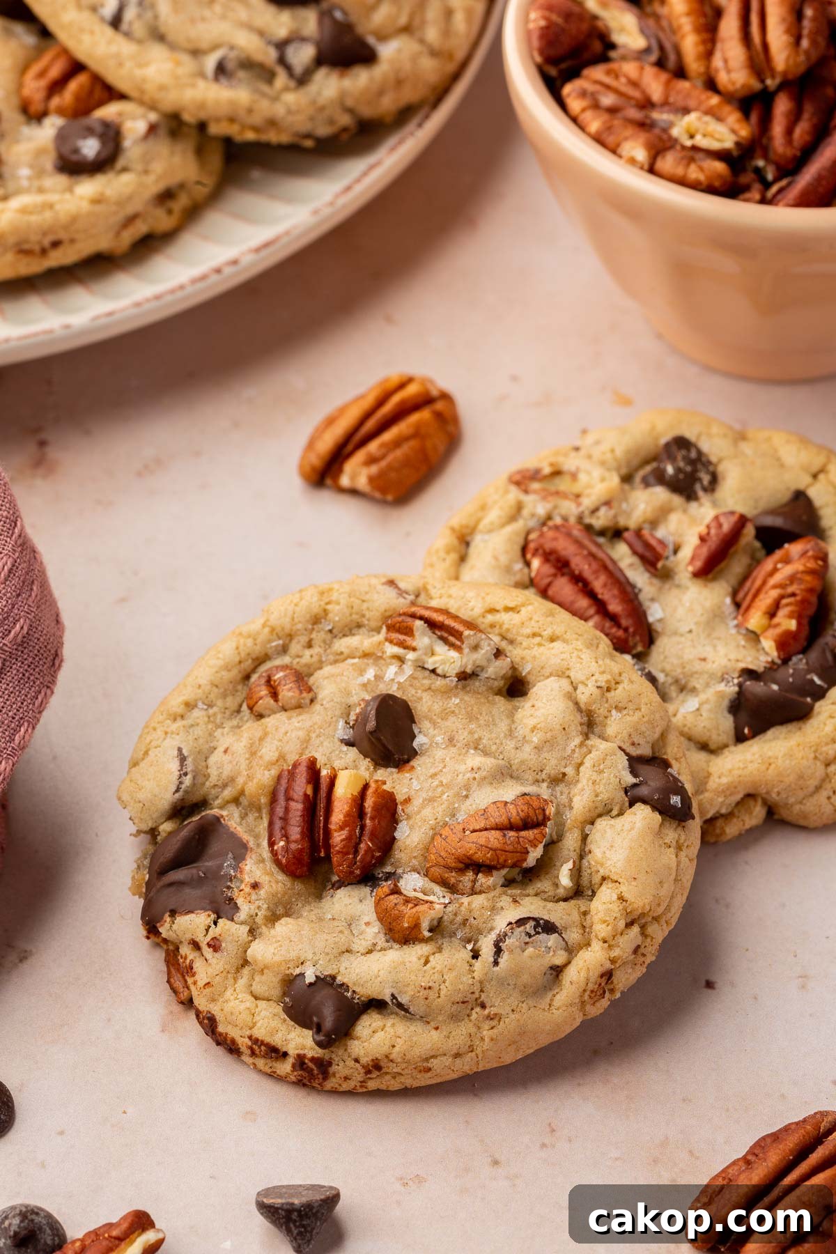 Two gluten free pecan chocolate chip cookies layered on top of each other on a pink table with additional gluten free cookies and toasted pecans in the background.