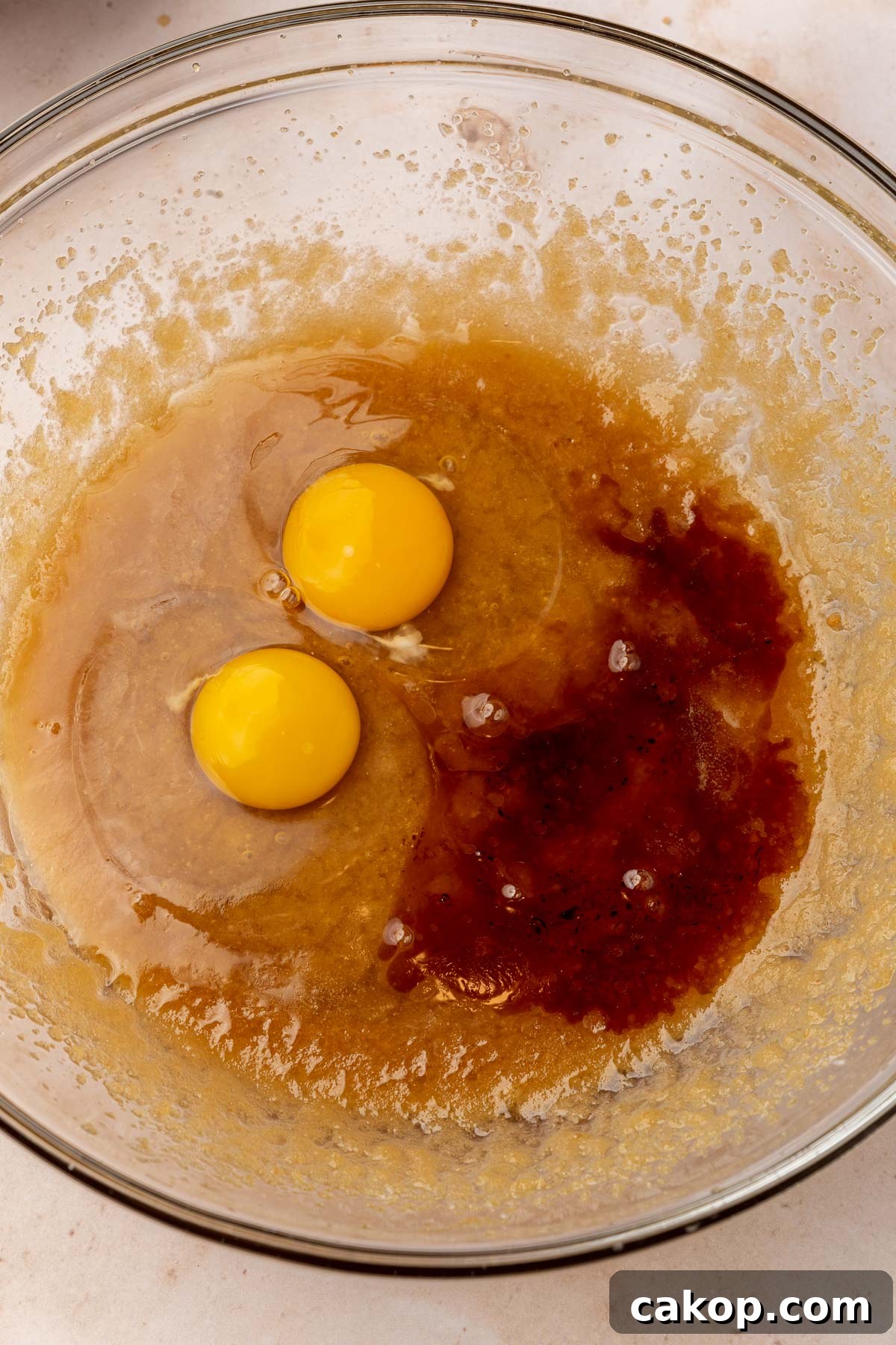 A glass mixing bowl with a mixture of melted butter, brown sugar and granulated sugar topped with pure vanilla extract and two eggs.