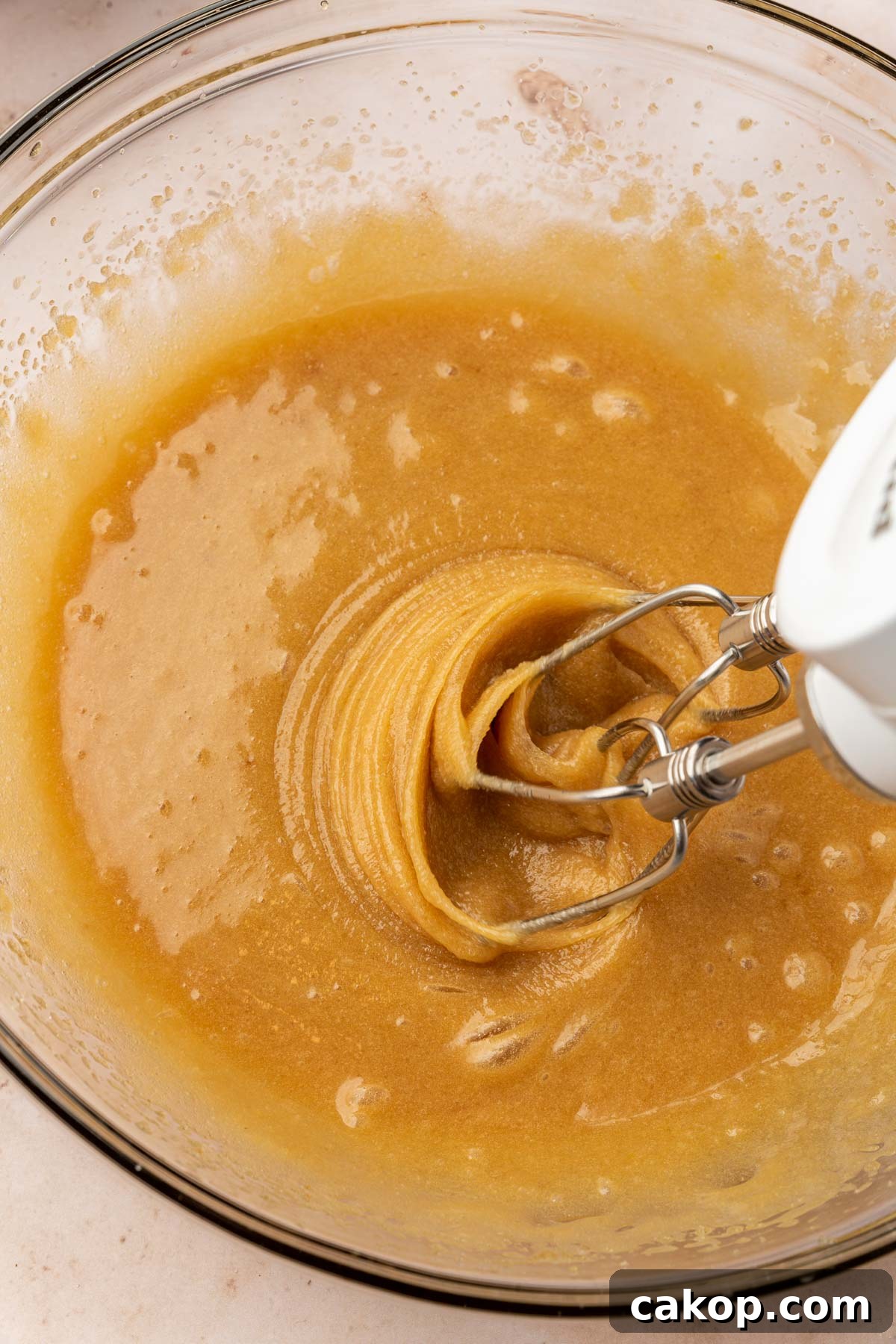 An electric mixer mixing together a cookie dough mixture of melted butter, brown sugar, sugar, vanilla, and eggs.