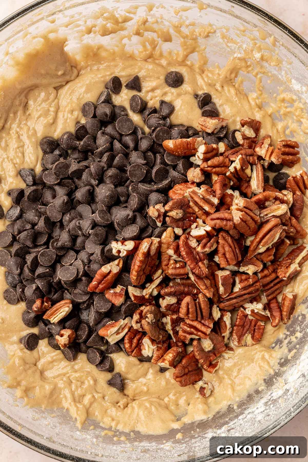 A close up of gluten free chocolate chip cookie dough topped with a pile of dark chocolate chips and toasted pecans before mixing together.