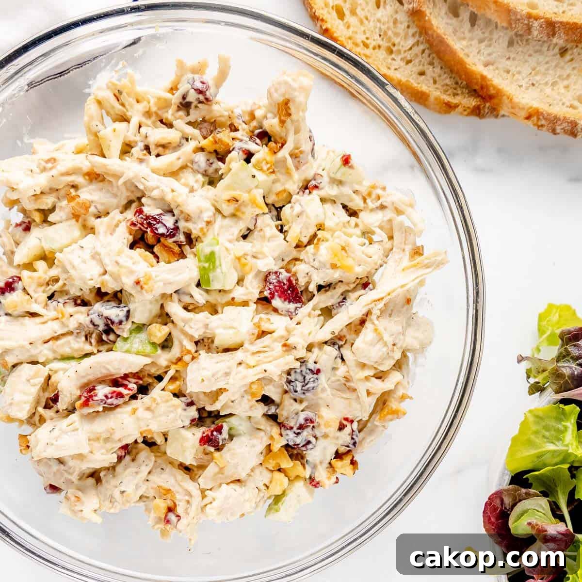 Classic Cape Cod Chicken Salad Recipe with Cranberries & Pecans 2 A glass bowl filled with chicken salad, featuring chopped chicken, cranberries, celery, walnuts, and creamy dressing. Accompanied by slices of bread and mixed greens on the side.