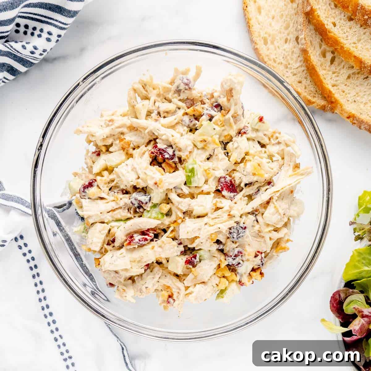 Classic Cape Cod Chicken Salad Recipe with Cranberries & Pecans 16 Glass bowl filled with chicken salad containing shredded chicken, grapes, nuts, and celery. Sliced bread and a striped cloth are nearby.