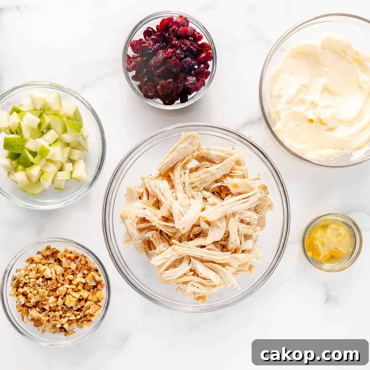 Classic Cape Cod Chicken Salad Recipe with Cranberries & Pecans 3 Ingredients for a salad on a white surface: shredded chicken, diced apples, mayonnaise, dried cranberries, chopped walnuts, and mustard in glass bowls.
