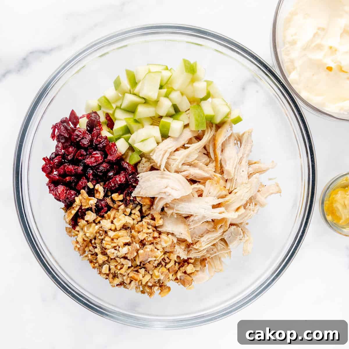 Classic Cape Cod Chicken Salad Recipe with Cranberries & Pecans 4 Glass bowl with diced green apples, shredded chicken, walnuts, and dried cranberries, set near a bowl of mayonnaise and a small dish of mustard on a marble surface.