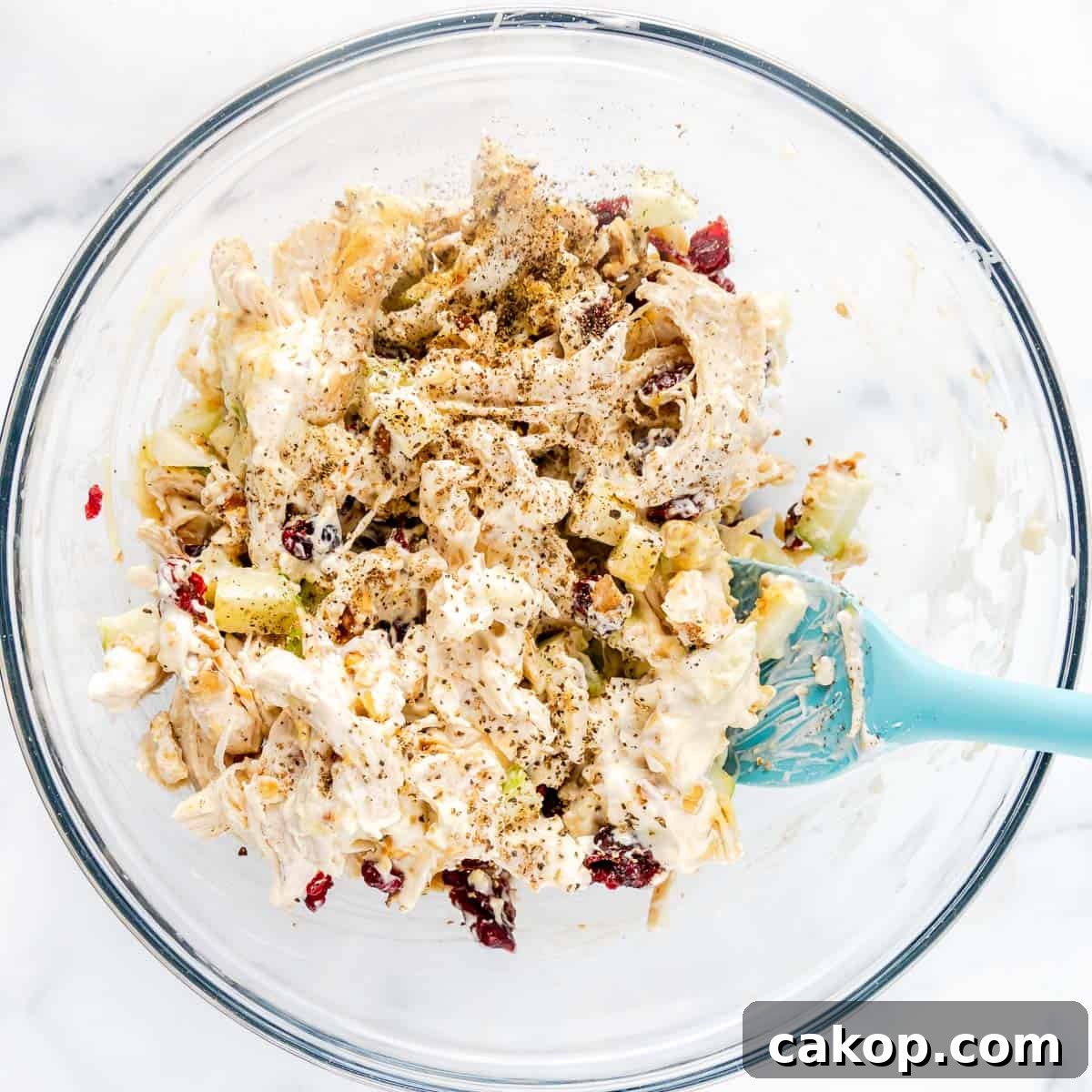 Classic Cape Cod Chicken Salad Recipe with Cranberries & Pecans 5 A glass bowl contains a mixture of shredded chicken, mayonnaise, nuts, celery, and cranberries, with a dusting of ground black pepper. A blue spatula is partially inserted in the mixture.