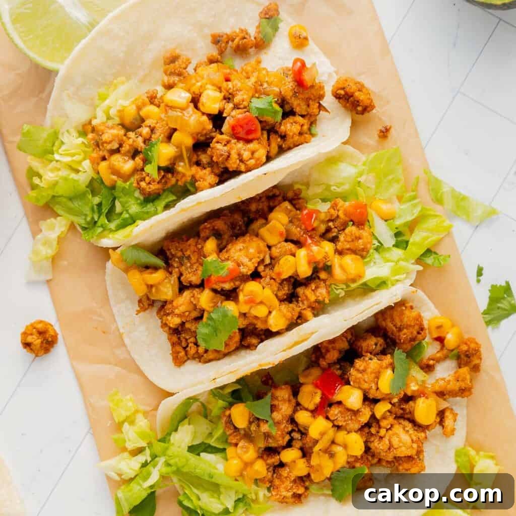 Three tacos filled with seasoned meat, corn, and lettuce on a parchment paper background. Lime wedges are partially visible in the corner.