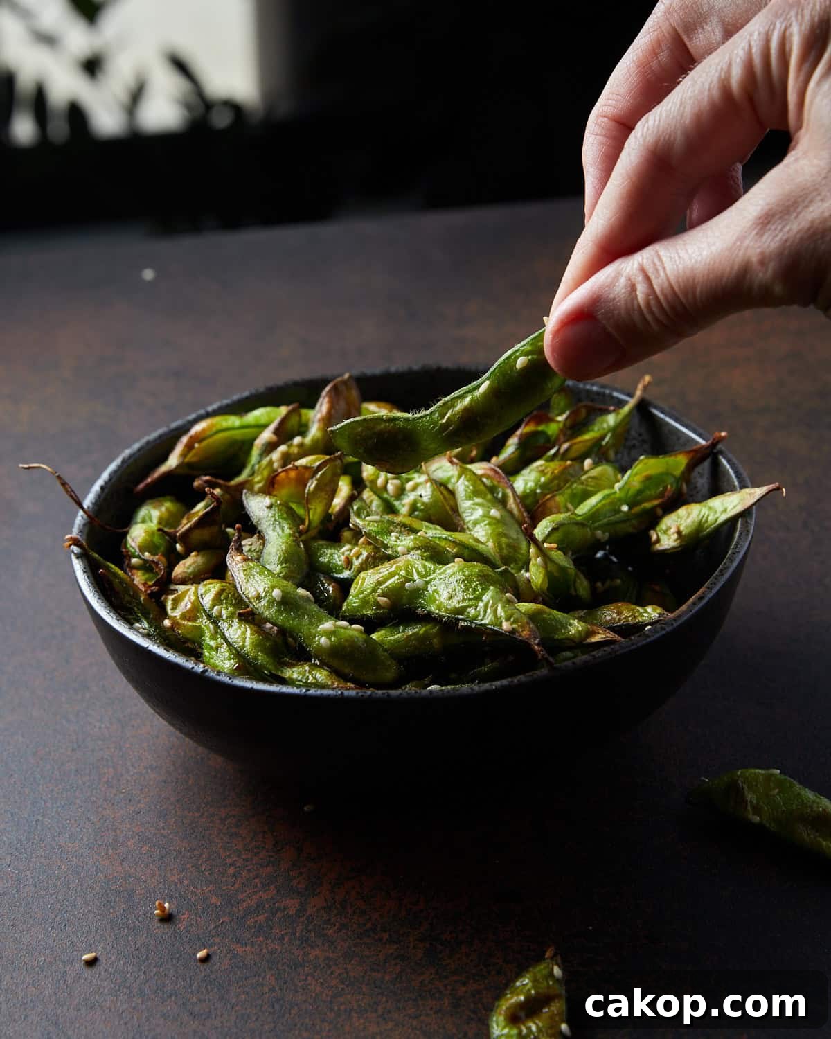 Crispy Air Fryer Frozen Edamame: Quick Snack or Side 5 Photo of a black bowl filled with air fryer frozen edamame beans set on a rusted black surface with a hand taking out one of the beans.