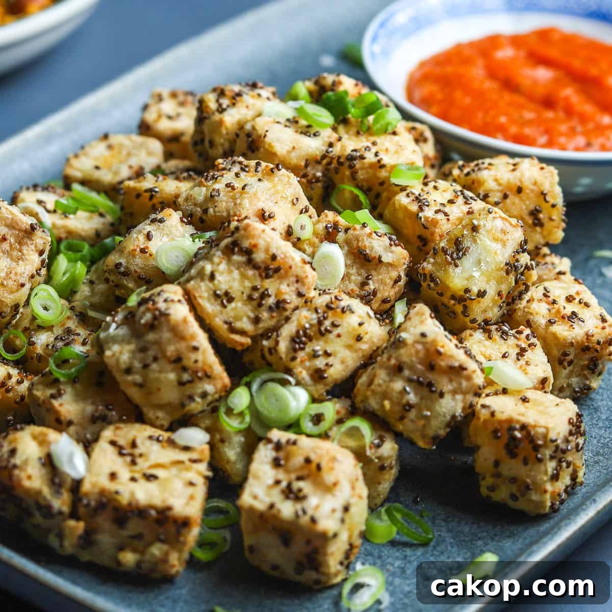 photo of a blue oblong plate filled with crispy air fried tofu with spring onion sprinkled on top and a bowl of kimchi sauce in the background.