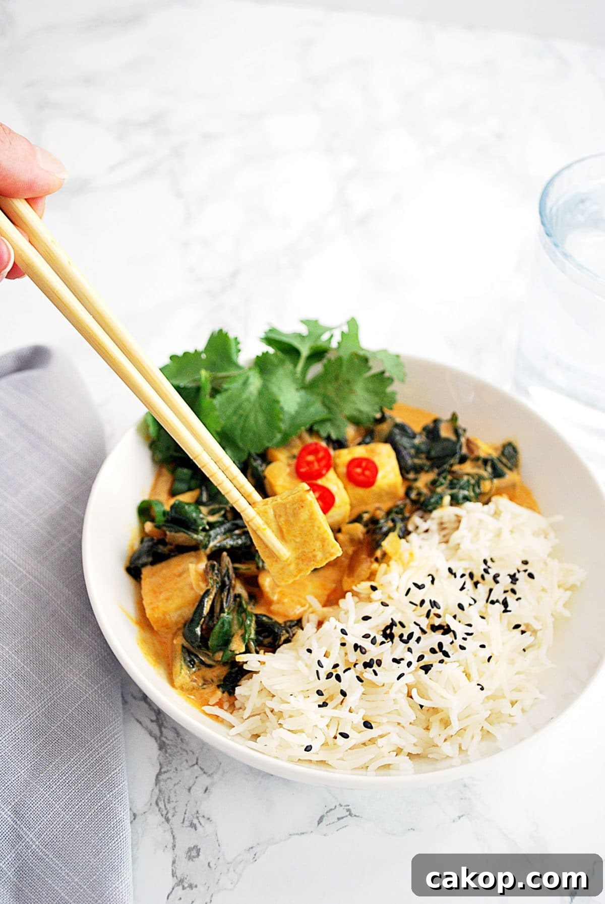 A piece of tofu from a bowl of red Thai curry tofu being held by chopsticks.
