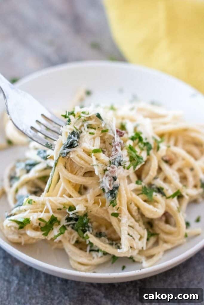Fork twirling creamy Tuscan pasta with greens and grated cheese on a white plate, placed on a gray table with a yellow napkin in the background.