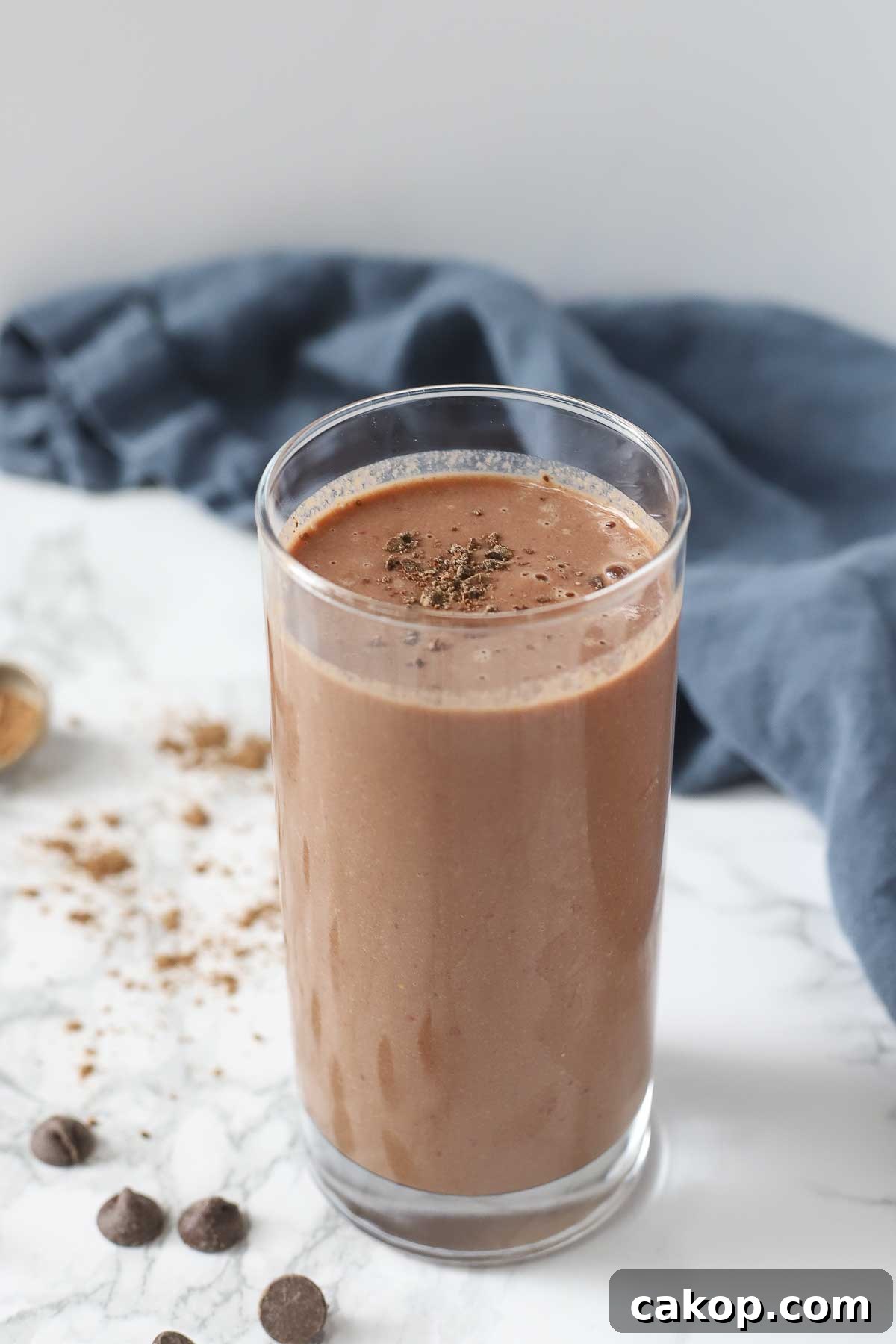 tall glass of chocolate cauliflower smoothie topped with chopped chocolate. The glass is on a white marble countertop and surrounded by chocolate chips, cocoa powder, and a blue napkin in the background.