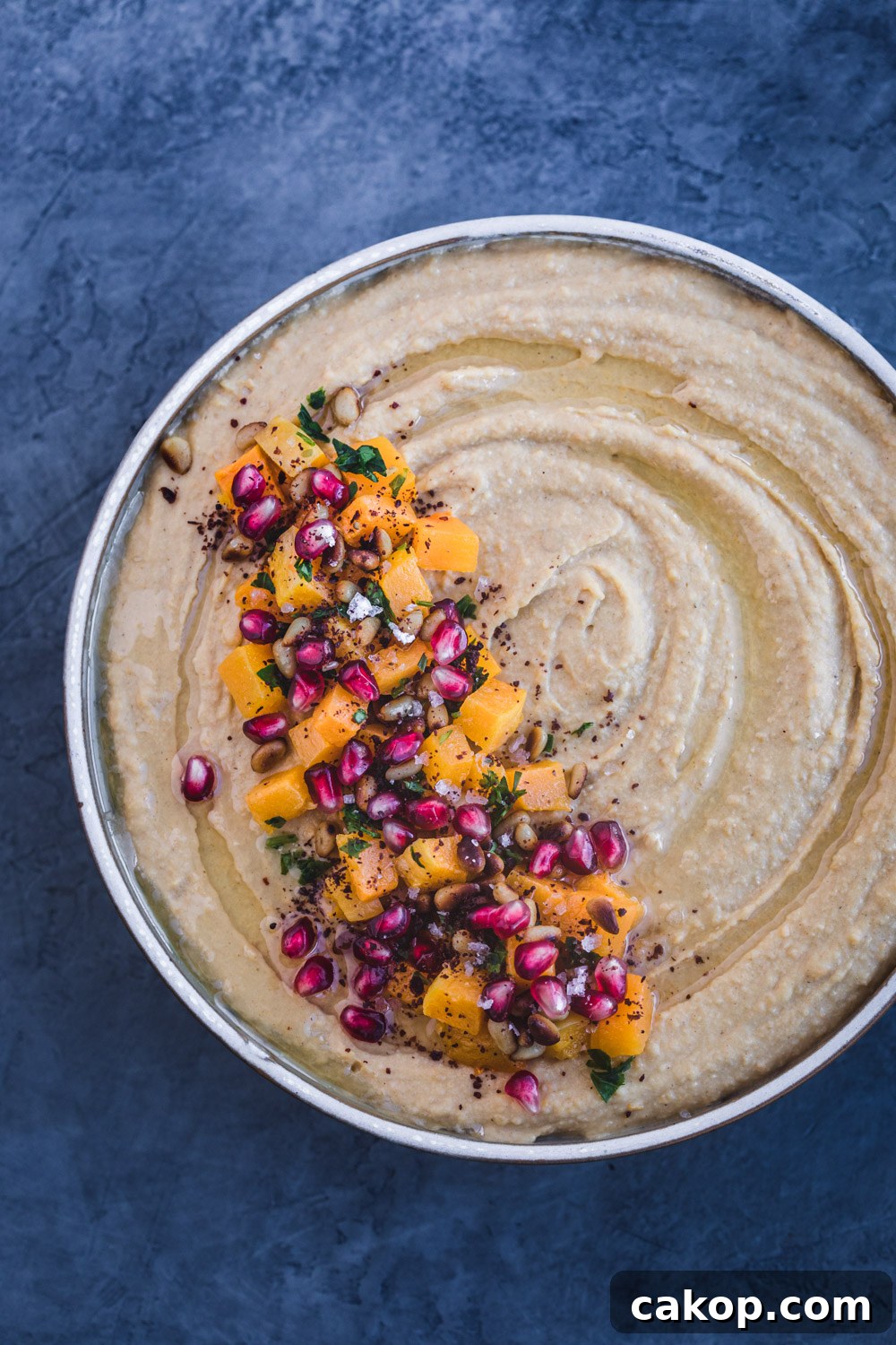 Overshot shot of butternut squash roasted hummus with cubed butternut roasted squash, pomegranate seeds, pastel, pinuts, sumac and flaky sea salt on top.