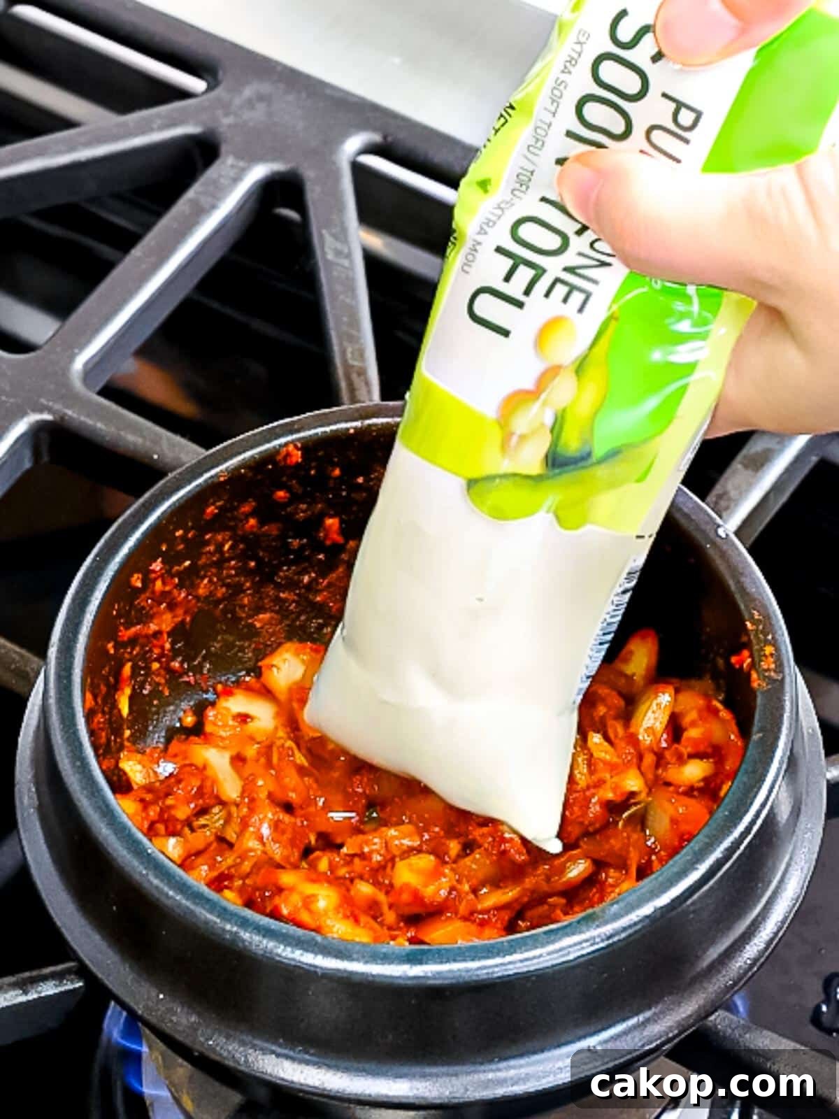 Spicy Korean Soondubu Jjigae (Silken Tofu Stew Recipe) 6 Korean soft tofu being added to kimchi in an earthenware pot.